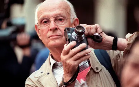 Un anziano fotografo francese famoso con occhiali tiene una vecchia macchina fotografica, pronto a catturare un momento.