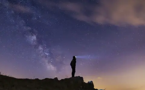 Silhouette di una persona su una collina, che guarda la Via Lattea. Un faro illumina il cielo stellato, un invito a scoprire la Via Lattea.