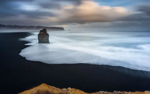 Scogliera solitaria su spiaggia nera, onde bianche in movimento con messa a fuoco infinito. Cielo drammatico.