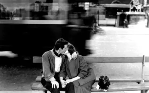 Un bacio rubato tra le opere di Robert Doisneau, un momento sospeso nel tempo con un tram sfocato sullo sfondo.