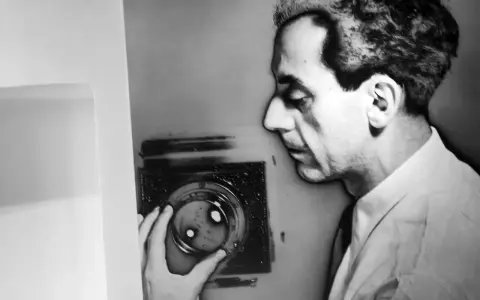 Man Ray, con la sua mano, manipola un oggetto che ricorda i suoi celebri rayogrammi, creando un'ombra enigmatica.