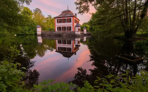 Castello bianco e arancione con torrette, riflessi nell'acqua calma al tramonto, circondato da alberi verdi.