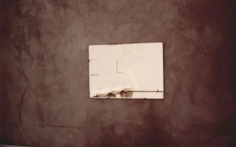 Specchio riflette lavandini in un bagno spoglio, un'immagine che evoca la poetica di Luigi Ghirri.