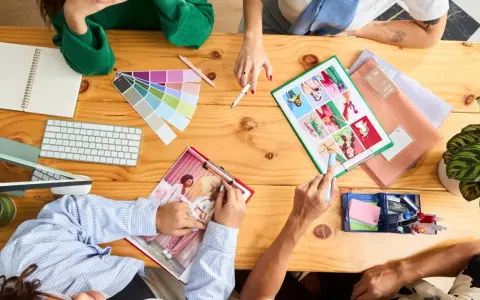 Un team creativo discute idee su un tavolo di legno, con campioni di colore, tastiera, quaderno e foto. Questo è un esempio di come una foto formato JPG può catturare un momento di collaborazione.