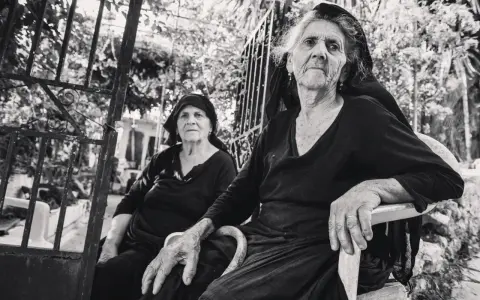 Due anziane donne, ritratte con la sensibilità di Paul Strand, siedono all'aperto, con sguardi intensi e mani segnate dal tempo.