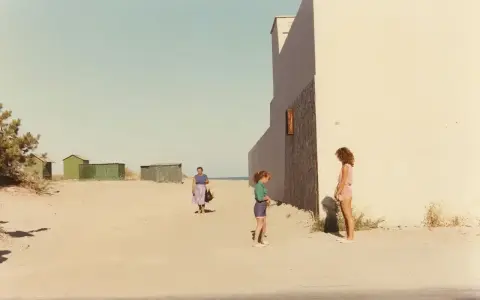 Ghirri fotografo cattura un momento estivo: due bambine e una donna sulla spiaggia, con capanne verdi sullo sfondo e un muro bianco imponente.