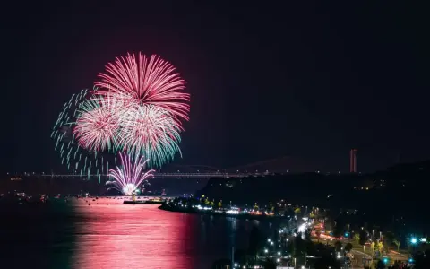 Spettacolare foto fuochi d'artificio rossi e verdi illuminano il cielo notturno sopra un fiume e una città con ponti illuminati.
