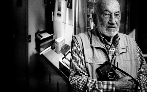 Ritratto di Gianni Berengo Gardin, con la sua inseparabile macchina fotografica al collo, sguardo intenso.