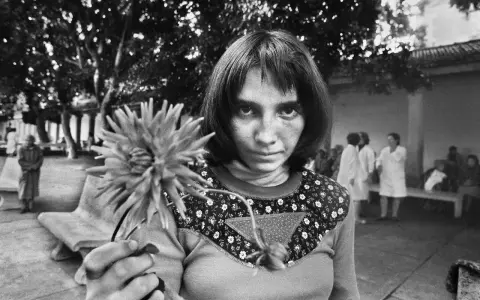 Letizia Battaglia giovane, con uno sguardo intenso, tiene un fiore. Sullo sfondo, figure sfocate in un cortile.