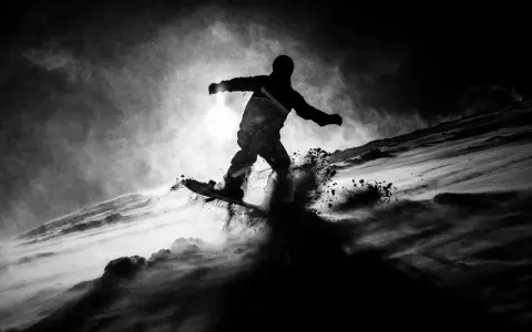 Snowboarder in silhouette, sollevando neve in una foto con ombre drammatiche.