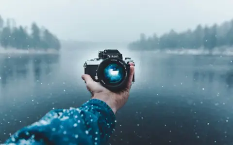 Mano che tiene una fotocamera Nikon in una giornata nevosa, con un lago e alberi sfocati sullo sfondo. Una foto raw per catturare l'essenza dell'inverno.