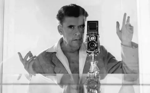 Herbert List si scatta un autoritratto con la sua Rolleiflex, un'immagine iconica che cattura il suo sguardo intenso e le mani in gesto.