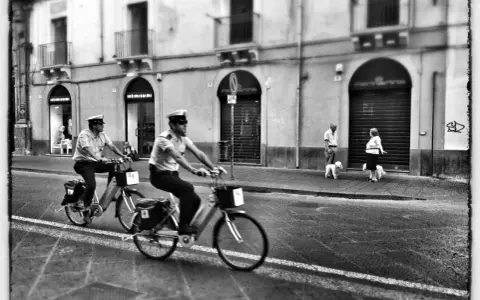 Due agenti in bicicletta pattugliano una strada italiana, mentre una coppia passeggia con i cani sul marciapiede. Italia street photography.