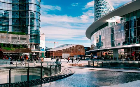 Un'area urbana moderna con grattacieli di vetro, un edificio curvo con il logo IBM e persone che passeggiano. Street photography esempi di vita cittadina.