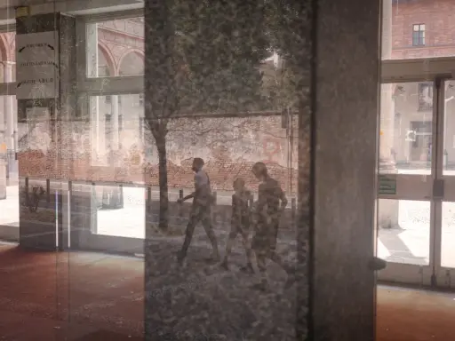 Un riflesso su vetro mostra tre figure che camminano, con un muro di mattoni e un edificio sullo sfondo.