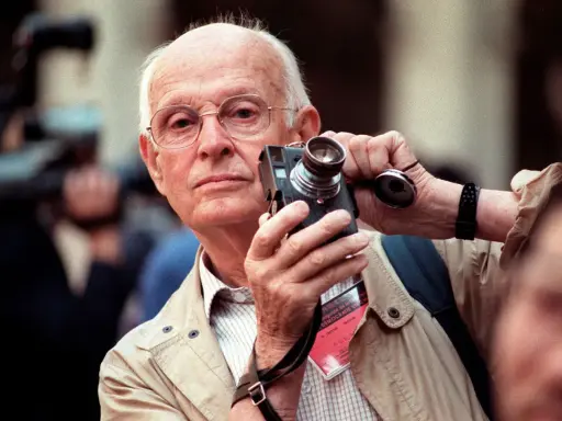 Un anziano fotografo francese famoso con occhiali tiene una vecchia macchina fotografica, pronto a catturare un momento.