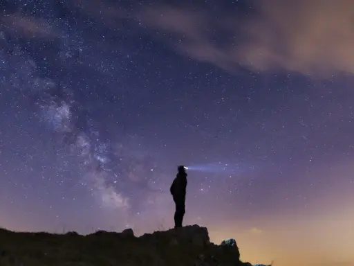 Silhouette di una persona su una collina, che guarda la Via Lattea. Un faro illumina il cielo stellato, un invito a scoprire la Via Lattea.