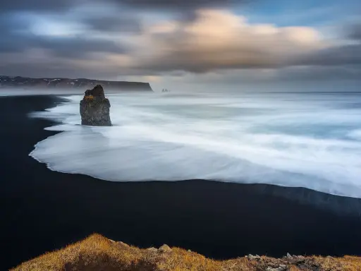 Scogliera solitaria su spiaggia nera, onde bianche in movimento con messa a fuoco infinito. Cielo drammatico.
