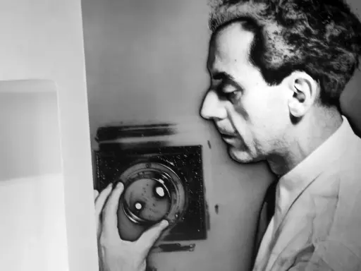 Man Ray, con la sua mano, manipola un oggetto che ricorda i suoi celebri rayogrammi, creando un'ombra enigmatica.