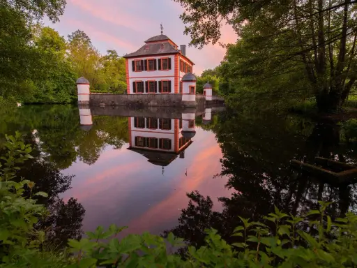 Castello bianco e arancione con torrette, riflessi nell'acqua calma al tramonto, circondato da alberi verdi.