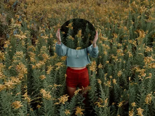 Una persona in un campo di fiori gialli tiene uno specchio rotondo davanti al viso, creando una **fotografia surrealista** dove il riflesso mostra solo la vegetazione circostante.