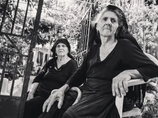 Due anziane donne, ritratte con la sensibilità di Paul Strand, siedono all'aperto, con sguardi intensi e mani segnate dal tempo.