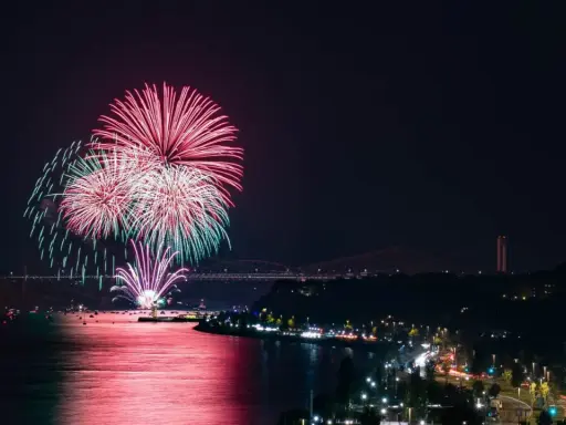 Spettacolare foto fuochi d'artificio rossi e verdi illuminano il cielo notturno sopra un fiume e una città con ponti illuminati.