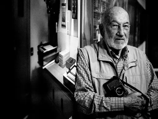 Ritratto di Gianni Berengo Gardin, con la sua inseparabile macchina fotografica al collo, sguardo intenso.