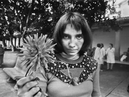 Letizia Battaglia giovane, con uno sguardo intenso, tiene un fiore. Sullo sfondo, figure sfocate in un cortile.