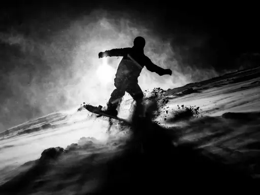 Snowboarder in silhouette, sollevando neve in una foto con ombre drammatiche.