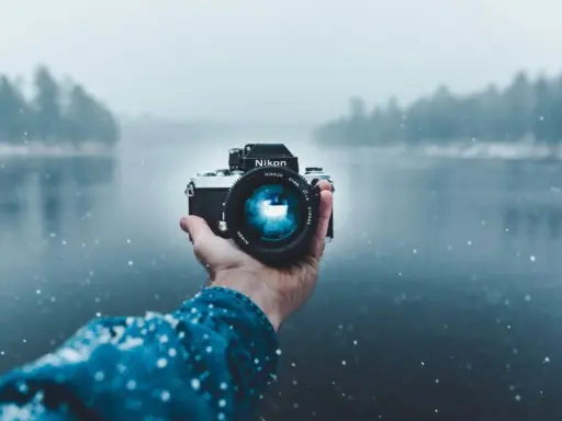 Mano che tiene una fotocamera Nikon in una giornata nevosa, con un lago e alberi sfocati sullo sfondo. Una foto raw per catturare l'essenza dell'inverno.