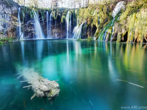 Cascata con effetto grandangolo, acqua turchese e un tronco sommerso.