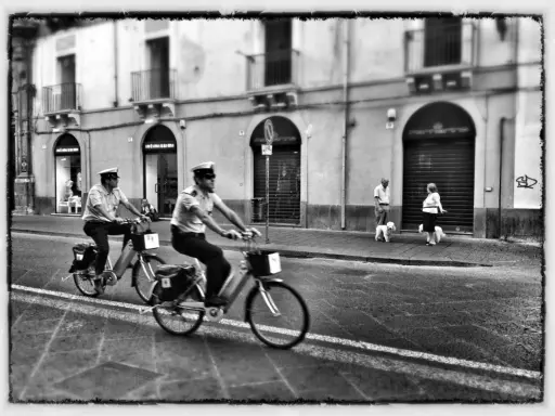 Due agenti in bicicletta pattugliano una strada italiana, mentre una coppia passeggia con i cani sul marciapiede. Italia street photography.