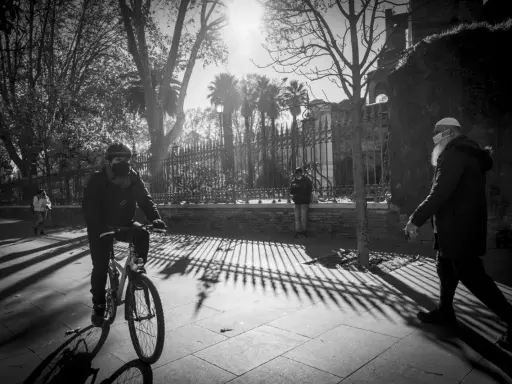 Ombre lunghe e un ciclista mascherato, in uno stile che ricorda robert doisneau, passeggiano sotto il sole.