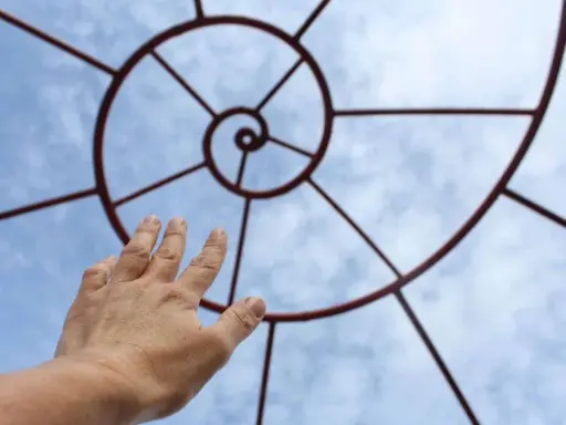 Mano tesa verso una spirale metallica che ricorda la composizione aurea, contro un cielo azzurro con nuvole.