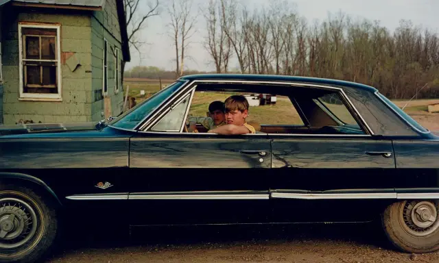 William Eggleston foto: due ragazzi guardano fuori da un'auto nera lucida, con una casa verde sullo sfondo.