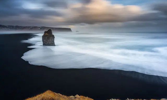 Scogliera solitaria su spiaggia nera, onde bianche in movimento con messa a fuoco infinito. Cielo drammatico.