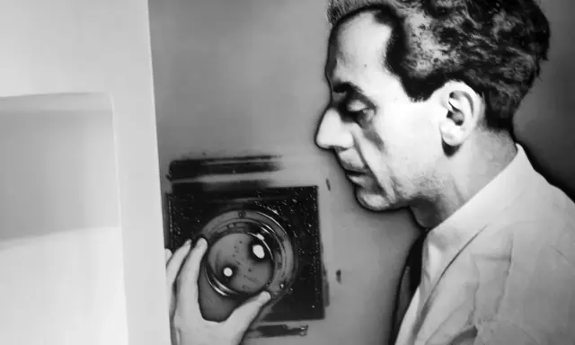 Man Ray, con la sua mano, manipola un oggetto che ricorda i suoi celebri rayogrammi, creando un'ombra enigmatica.