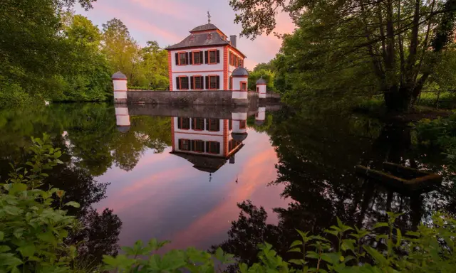 Castello bianco e arancione con torrette, riflessi nell'acqua calma al tramonto, circondato da alberi verdi.