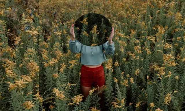 Una persona in un campo di fiori gialli tiene uno specchio rotondo davanti al viso, creando una **fotografia surrealista** dove il riflesso mostra solo la vegetazione circostante.