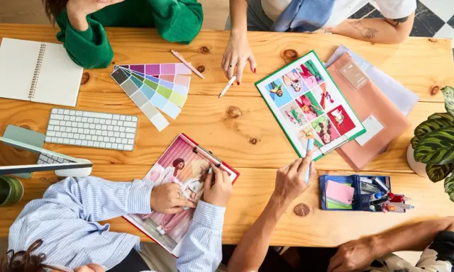 Un team creativo discute idee su un tavolo di legno, con campioni di colore, tastiera, quaderno e foto. Questo è un esempio di come una foto formato JPG può catturare un momento di collaborazione.