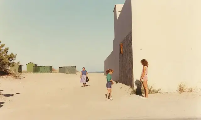 Ghirri fotografo cattura un momento estivo: due bambine e una donna sulla spiaggia, con capanne verdi sullo sfondo e un muro bianco imponente.