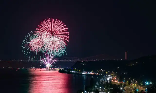 Spettacolare foto fuochi d'artificio rossi e verdi illuminano il cielo notturno sopra un fiume e una città con ponti illuminati.