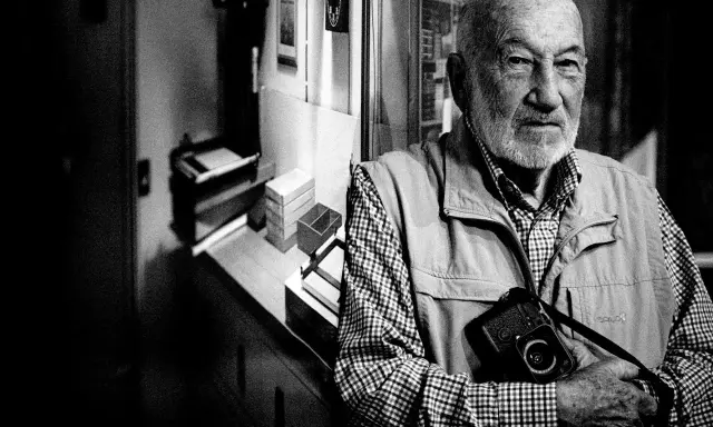 Ritratto di Gianni Berengo Gardin, con la sua inseparabile macchina fotografica al collo, sguardo intenso.