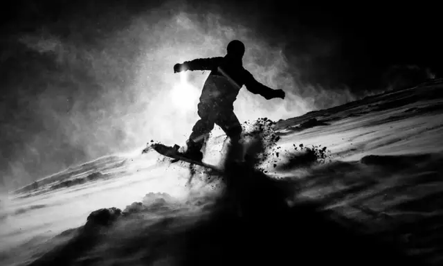 Snowboarder in silhouette, sollevando neve in una foto con ombre drammatiche.