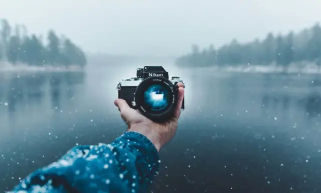 Mano che tiene una fotocamera Nikon in una giornata nevosa, con un lago e alberi sfocati sullo sfondo. Una foto raw per catturare l'essenza dell'inverno.