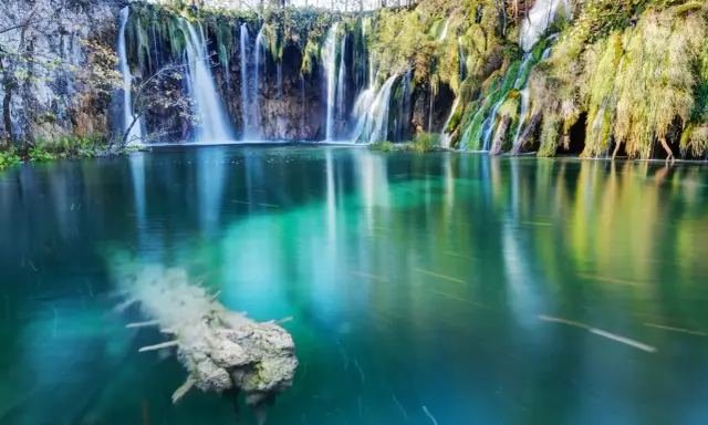 Cascata con effetto grandangolo, acqua turchese e un tronco sommerso.