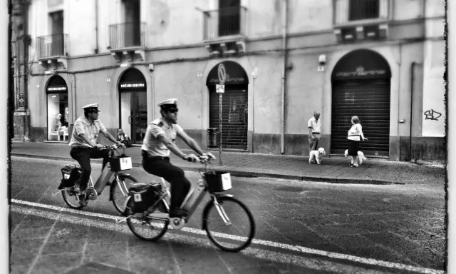 Due agenti in bicicletta pattugliano una strada italiana, mentre una coppia passeggia con i cani sul marciapiede. Italia street photography.