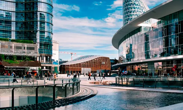 Un'area urbana moderna con grattacieli di vetro, un edificio curvo con il logo IBM e persone che passeggiano. Street photography esempi di vita cittadina.