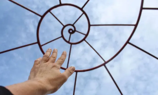 Mano tesa verso una spirale metallica che ricorda la composizione aurea, contro un cielo azzurro con nuvole.